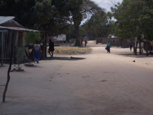 Die Hauptstraße des Dorfes mit einigen Läden am Straßenrand. Im Hintergrund ist ein großes Werbeplakat für Kondome zu sehen.